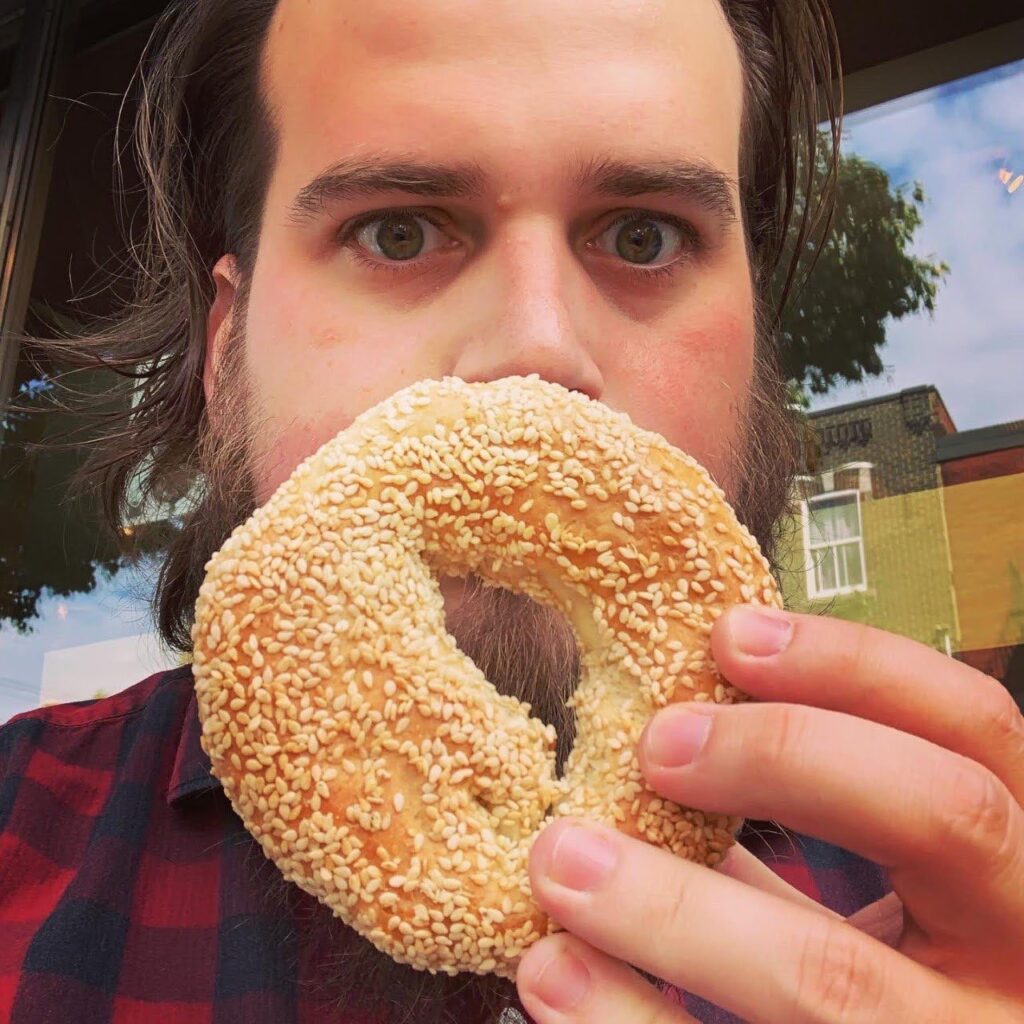 Robert Peiffle holding a sesame bagel in front of his face like a picture frame, standing in front of a window with a tree and building reflected in it.
